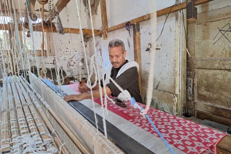 63 سنة ولسة بيعافر.. شيخ النساجين في سوهاج: عملت تحفة فنية على قماشة مدهبة والسر في النول اليدوي على آلة خشبية معقدة بها خيوط ملونة وقد تكون بيضاء فقط، ينسج العم عبد الصبور، شيخ ال